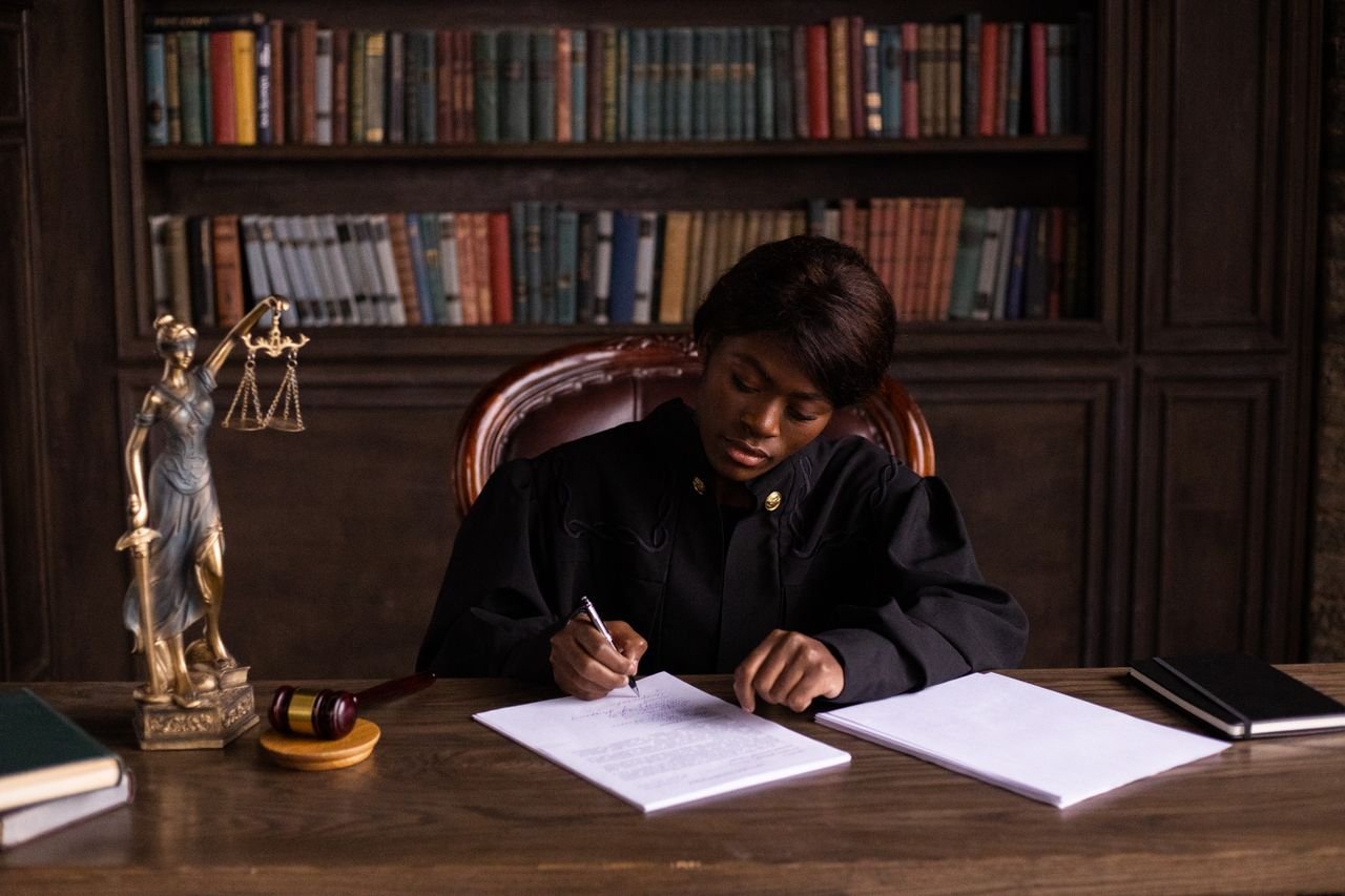 about-01 A judge in robes writing on a document at a desk in an office library with law books.